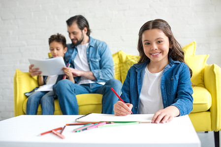 孩子彩色铅笔摄影照片_可爱的孩子绘图与彩色铅笔和微笑的相机, 而父亲和弟弟坐在沙发后面 
