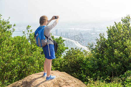年轻的女性游客拍照, 欣赏风景, 清新的空气和风在山顶上的绿色灌木。女孩与智能手机和蓝色背包。海滨城市在背景下可见