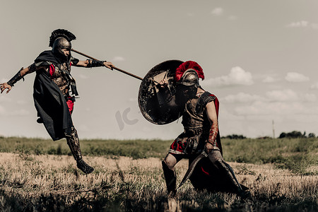 沙场战士摄影照片_两个身穿战斗服的古希腊战士或罗马战士，在天空的衬托下，在草地上，用矛和剑战斗.