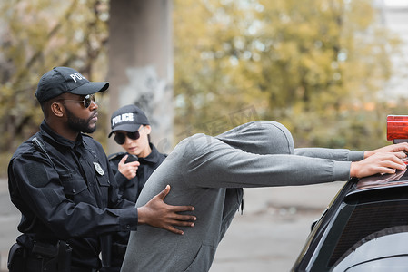 蒙面舞会男女摄影照片_非洲裔美国警察搜刮蒙面罪犯，靠在同事身旁的巡逻车上，在室外模糊的背景下