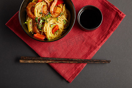 筷子上的面摄影照片_在木制筷子旁边的碗里放些虾仁和蔬菜的面，在黑色背景的红餐巾纸上放些酱油