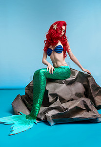 在蓝色背景上坐在石头上的红色波浪状头发的美人鱼