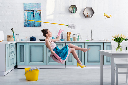 优雅的女孩在围裙悬浮与拖把和黄色的鞋跟鞋在厨房的房子清洁