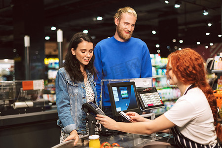年轻的微笑夫妇站在收银台附近愉快地支付在现代超市的信用卡产品