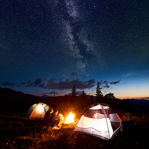 家庭徒步旅行的母亲, 父亲, 两个儿子晚上在山上露营, 坐在篝火旁的日志和两个照明帐篷, 在令人惊叹的夜晚天空充满星星, 银河