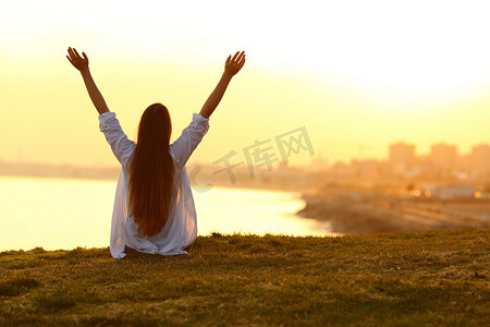 坐在草地摄影照片_后视图的照片一个快乐的女人举起手臂坐在草地上日落与沿海城市的背景