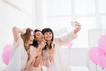 睡衣美女摄影照片_睡衣派对上，带着香槟酒的多文化美女在智能手机上自拍