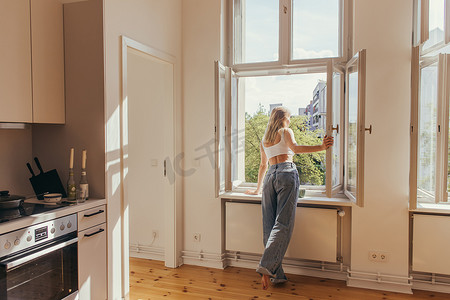 金发女人看着厨房窗台上靠近一杯咖啡的开着的窗户 