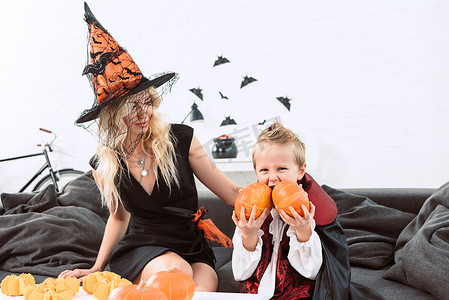 在吸血鬼万圣节服装小男孩的肖像咬南瓜坐在沙发上与母亲在巫婆服装
