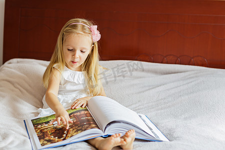 可爱小孩坐摄影照片_可爱的小女孩穿着时髦的白色衣服看书。小孩坐在房间的床上。在coronavirus covid-19封锁期间呆在家里