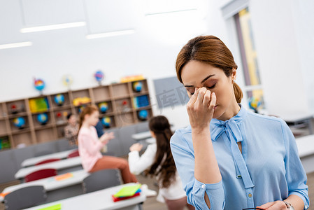穿着蓝色上衣的疲惫的老师站在课桌前, 摸着鼻子
