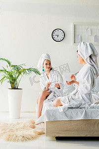 穿着浴袍和珠宝的时尚女性，头上的毛巾坐在床上，在绿色植物附近说话和钉指甲，在地板上的毛皮
