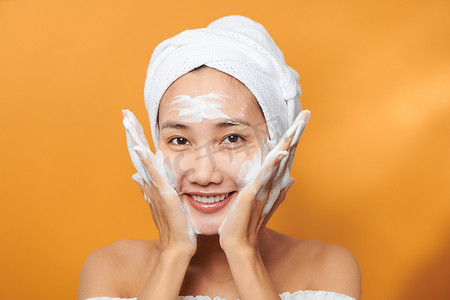 Happy young Asian woman applying face cream while wearing a towel and touching her face. Isolated on orange background