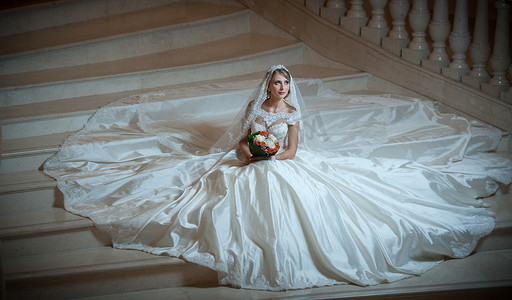 年轻漂亮的豪华女人穿着婚纱，在半黑暗中坐在楼梯台阶上。抱着她的花束的巨大婚纱的新娘。诱人的金发新娘，在袭华美的袍摆姿势