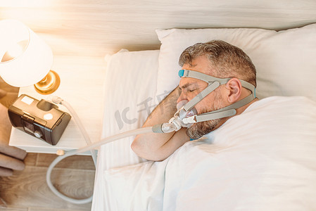 考虑考虑摄影照片_有慢性呼吸问题的睡眠男子考虑在床上使用CPAP机。保健、阻塞性睡眠呼吸暂停疗法、 CPAP 、打鼾概念