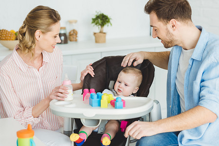 婴儿与奶瓶摄影照片_父母与婴儿的女儿坐在婴儿椅与塑料块和奶瓶