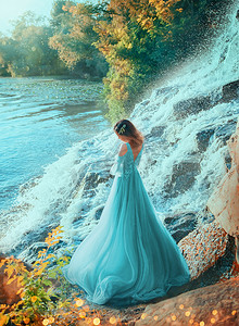 小精灵女孩站在岸边的湖上，转身离去。石头溅水。金色的夏秋红头发收集装饰过的花。蓝色绿松石晚长而茂密的连衣裙。毕业舞会艺术