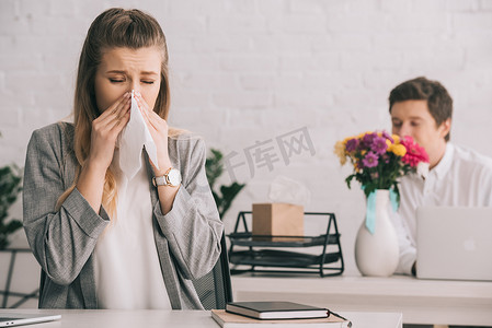 嗅到摄影照片_金发碧眼的女商人与花粉过敏打喷嚏在组织附近的同事嗅到鲜花在办公室 