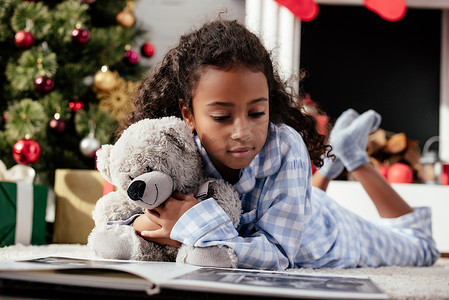 pajamas摄影照片_adorable african american child in pajamas with teddy bear looking at photo album on floor at home