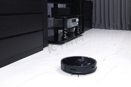 漂亮的黑色现代自动扫描机器人真空地带正在把漂亮的卧室里肮脏的大理石地板劈开，房间里有橱柜、电视机和灰色窗帘