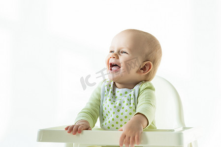 Cheerful baby in bib looking away on feeding chair on white background