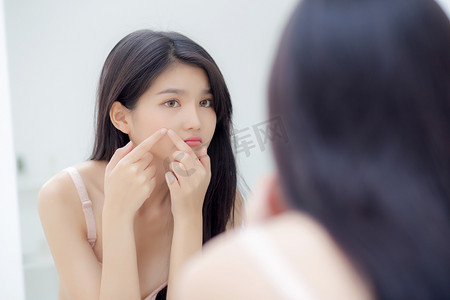青春是美丽的摄影照片_美丽的年轻亚洲女人看着镜子和触摸的脸，是一种痤疮、青春痘、美容美发、脸上皱纹、化妆品和化妆品的概念.