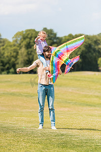 快乐的父亲背着小女儿在脖子上玩五颜六色的风筝在公园里 