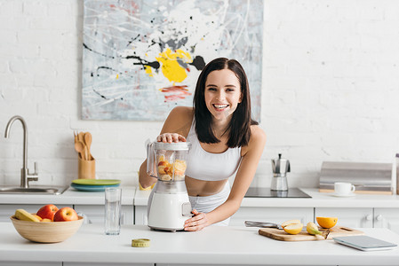 Selective focus of smiling sportswoman preparing smoothie near measuring tape and fruits on kitchen table, calorie counting diet