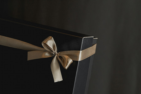 气氛金色摄影照片_黑色礼品盒的特写镜头，带着优雅的金色缎带，背景是深色的豪华蝴蝶结。带有文字位置的礼品盒。生日,结婚周年礼物,礼品明信片.浪漫的气氛