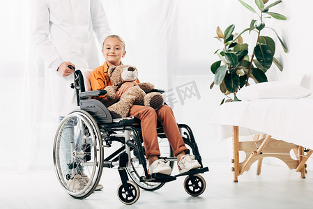 穿着白色外套的花哨和在轮椅上带着泰迪熊的孩子的裁剪视图
