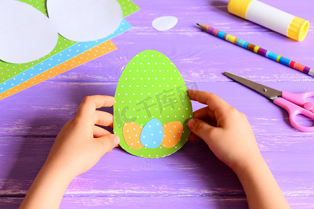 儿童剪纸活动摄影照片_小孩子手里拿着复活节纸卡.孩子们制作了有创意的彩蛋形复活节贺卡.在木制桌子上进行艺术活动的材料.幼稚园或家中手工造纸工艺概念