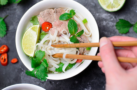 酸辣鸡胗米粉摄影照片_传统越南汤 pho bo 与米粉, 牛肉和草药在黑暗的背景, 亚洲美食, 在女性手的筷子