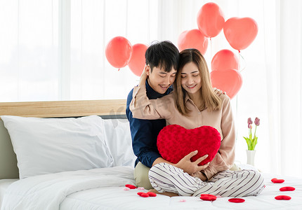 垫子摄影照片_乐观的一对年轻的亚洲夫妇，带着心形的垫子，一边微笑一边放松，在床上拥抱对方。爱与青少年恋爱关系的概念.