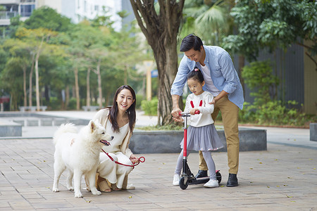 亚洲父母和女儿玩滑板车, 而遛狗在花园
