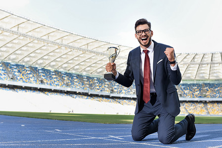 身穿西服的快乐的年轻商人跪在跑道上，在体育馆里拿着奖杯
