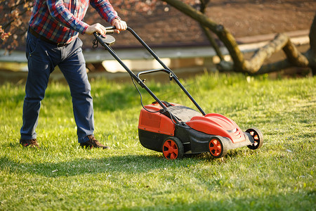 割摄影照片_日落时分，割草工人在绿色的院子里割草。带着电动割草机割草坪的人园艺师修剪花园.