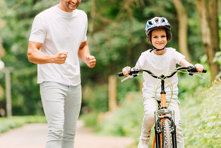 男孩骑摄影照片_裁剪视图的父亲欢呼的儿子，而男孩骑自行车，看着相机