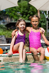 水拥抱摄影照片_穿着泳衣的孩子们坐在游泳池边，双腿在水里，拥抱着，看着镜头
