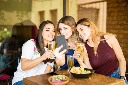 喝啤酒摄影照片_三名年轻女性坐在餐馆里喝啤酒, 带着自拍移动喷