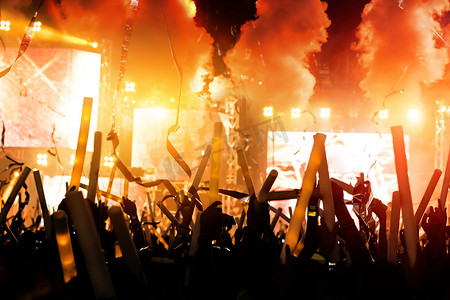 爆炸冲击效果摄影照片_音乐会舞台灯光的人群和人们球迷观众的剪影提高手或发光棒举行在音乐节后视镜与聚光灯发光的效果和烟雾