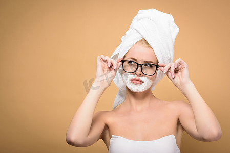 young woman with towel on head and shaving cream on face adjusting eyeglasses and looking away isolated on beige