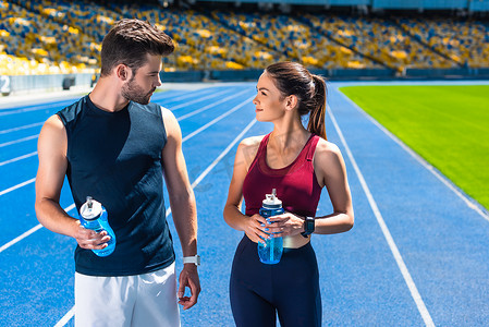 年轻的嬉戏夫妇与瓶装水聊天在体育场馆的运行轨道