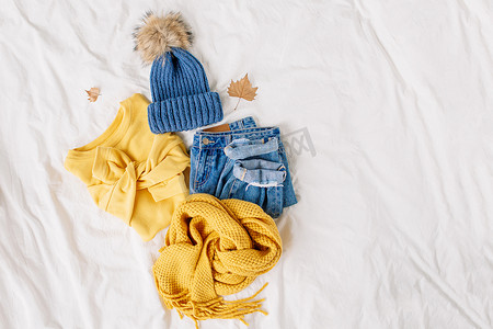 舒适的黄色毛衣，蓝色牛仔裤，围巾和帽子在床上的白床单。女士时尚的秋冬套装。时髦的服装拼贴画。平铺，顶视图. 