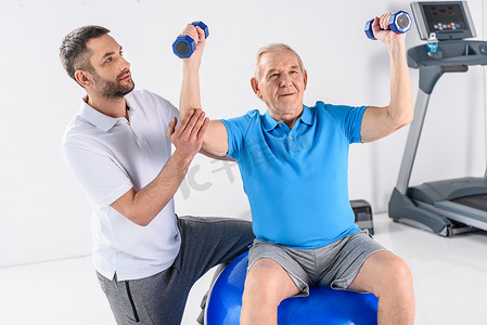 康复治疗师在灰色背景下协助在健身球上锻炼哑铃的老人