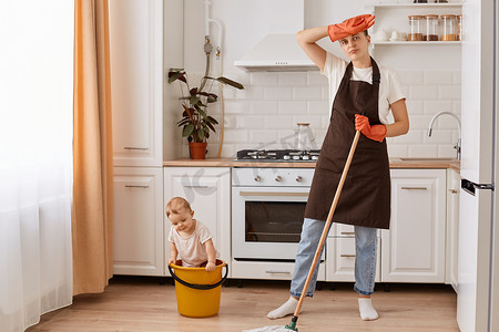 Portrait of tired woman washing floor, wiping her forehead at kitchen, female wearing brown apron, standing, looking at camera, her daughter playing in the bucket, helping mother with cleaning.