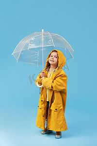 端午节快乐字体摄影照片_快乐有趣的孩子,在蓝色的工作室背景上摆出透明的伞.女孩穿着黄色雨衣和橡胶靴。持有一个老式旅行箱