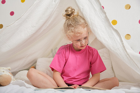 孩子们的房间摄影照片_一个头发卷曲的可爱女孩从床上的床单上读了一本书，小女孩在她的帐篷里玩得很开心，在孩子们的房间里打闹
