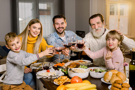 美味共享受摄影照片_家庭共进晚餐庆祝概念，感恩节假期。 一家人幸福地坐在桌旁笑着，享受着在一起的时光，享用着美味的晚餐，手里拿着酒杯和果汁