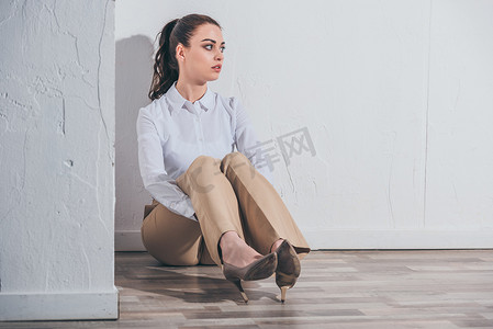 心烦意乱的女人在白色的衬衫和米色的裤子坐在地板上靠近白色墙壁在家里, 悲伤的混乱的概念