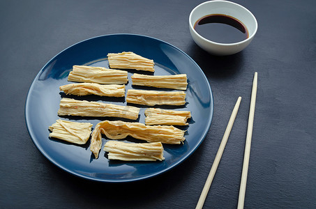 豆腐摄影照片_豆豆腐的皮在蓝色盘子里，用竹竿和酱油在黑色的板岩背景上。东亚菜。素食精益产品。有测试的地方